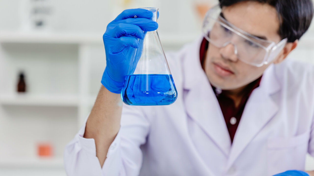 Junger Mann mit Schutzbrille und blauen Handschuhen, hält Glasbehälter mit blauer Flüssigkeit in der Hand