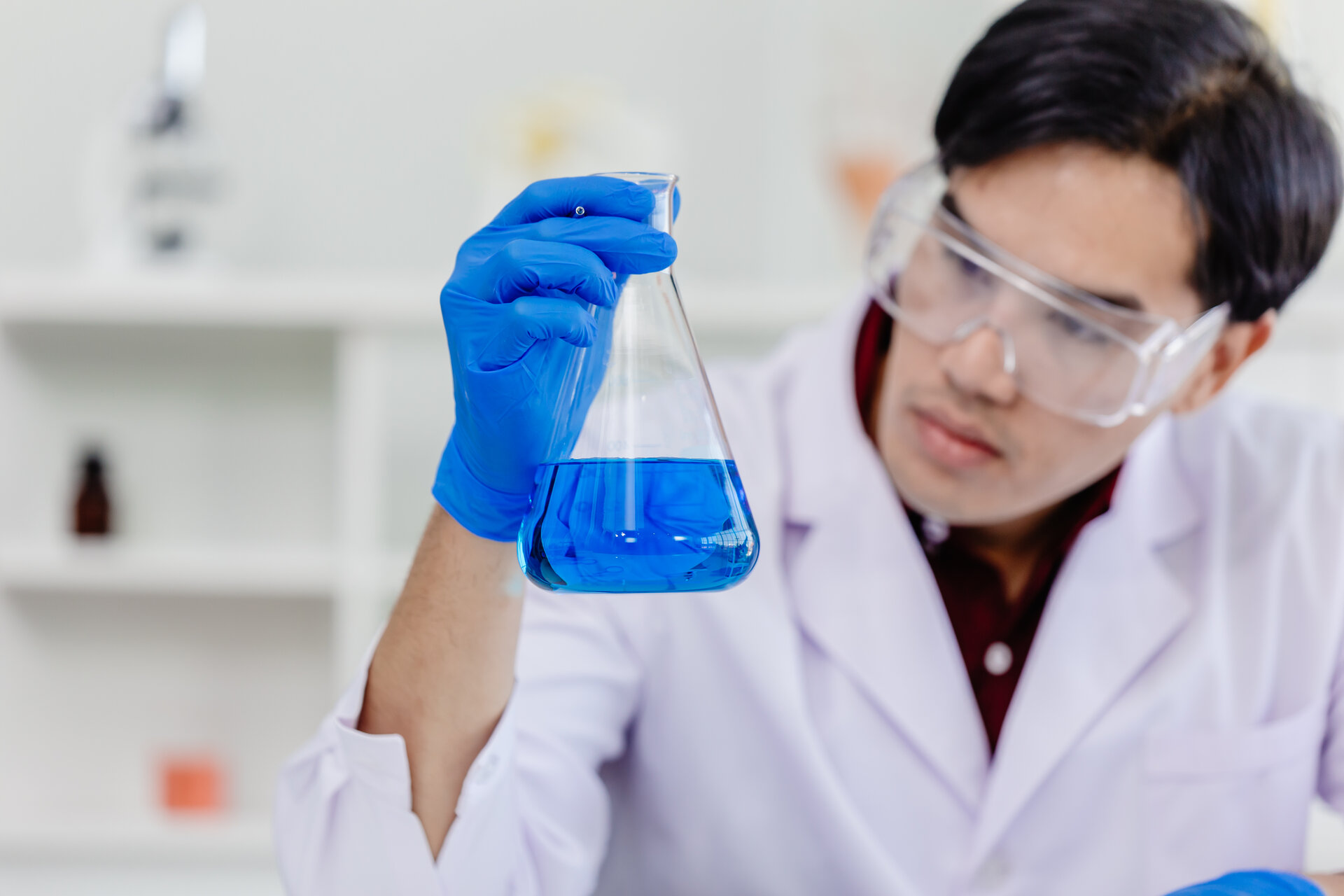 Junger Mann mit Schutzbrille und blauen Handschuhen, hält Glasbehälter mit blauer Flüssigkeit in der Hand