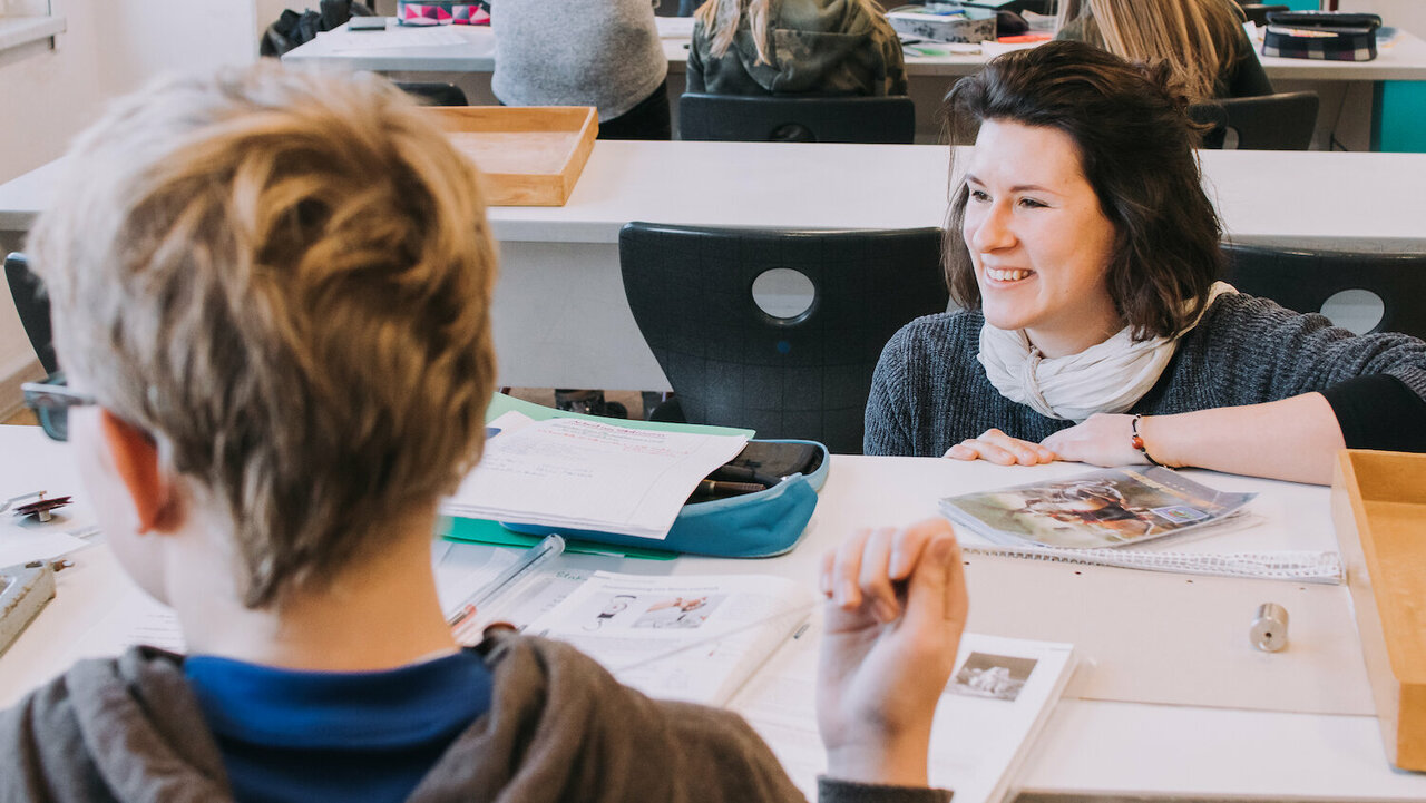 Blondes Kind von hinten, dunkelhaarige Lehrerin lehnt sich auf die Schulbank