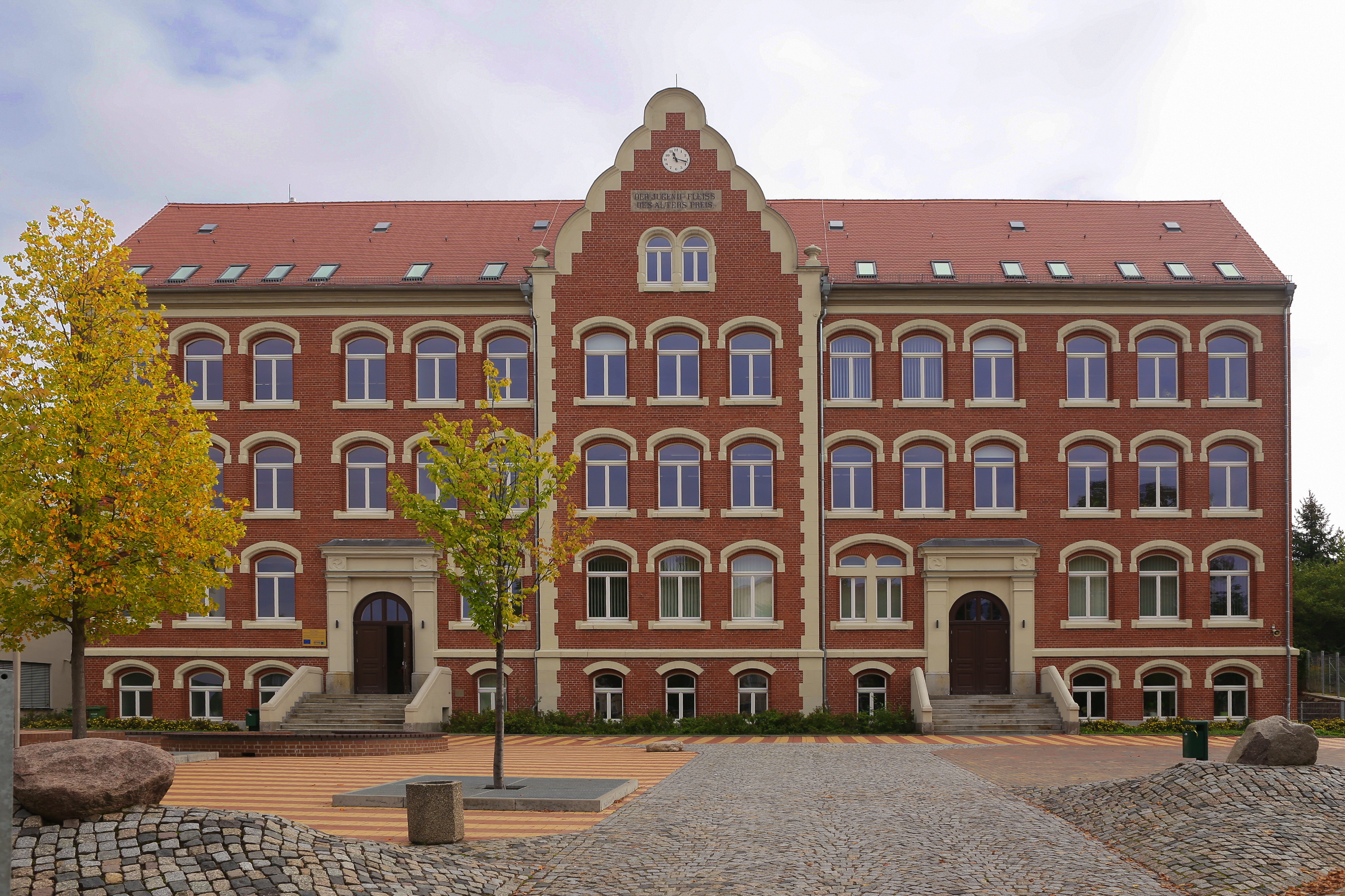 Vorderansicht des Schulgebäudes, Backsteinbau mit 3 Etagen und 2 Schuleingänge links und rechts, davor Teile des gepflasterten Schulhofes